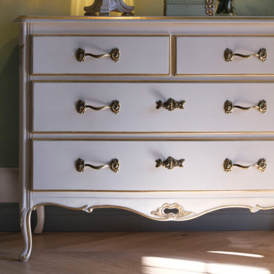Ornate Louis Style Chest Of Drawers An Ornate Louis Style Chest Of Drawers, featuring a vintage design with white finish and ornate gold accents, offers seven drawers for ample storage. The piece is elevated on elegantly curved legs. Sunlight casts a warm glow on its surface, creating a cozy ambiance against the soft green painted wall behind it.
