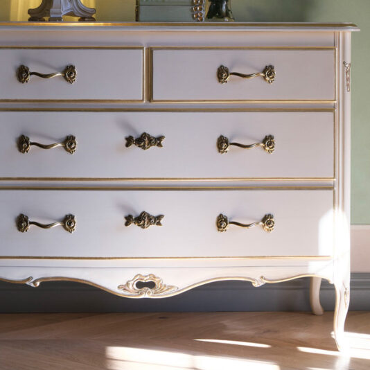 Ornate Louis Style Chest Of Drawers An Ornate Louis Style Chest Of Drawers with a vintage design, featuring gold handles and intricate detailing in white, sits against a light green wall. The dresser includes two small drawers on the top row and three larger drawers below. Sunlight casts shadows on the wooden floor in the foreground.