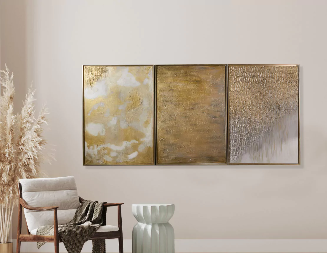 A minimalist living space, embodying Organic Modern Style, features a beige wall adorned with a triptych of abstract gold and white textured paintings. Nearby is a wooden armchair with a beige cushion and brown throw, beside a green ribbed side table. Dried pampas grass decorates the corner.