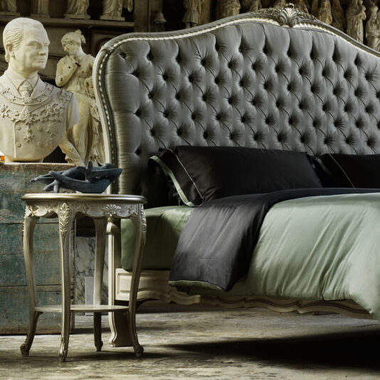 A luxurious bedroom showcases a tufted, silver headboard and bedding in rich shades of green and black. Beside the bed stands the Rococo Style Round Bedside Table, adorned with a bust and various decorative items. The room exudes a classic, opulent aesthetic complemented by statues in the background.