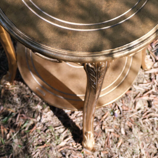 Rococo Style Round Gold Bedside Table A close-up of a Rococo Style Round Gold Bedside Table featuring ornate details and intricate carved legs. The table has two circular tiers and is positioned on a patch of ground covered with dry leaves and grass. Sunlight casts shadows on the surface.