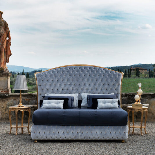Rococo Style Round Gold Bedside Table A luxurious, tufted bed with a tall, curved headboard stands outdoors on a gravel surface, flanked by two elegant Rococo Style Round Gold Bedside Tables each adorned with matching lamps. Beyond the bed, a scenic view of rolling green hills and a clear sky is visible. A statue is on the left side.
