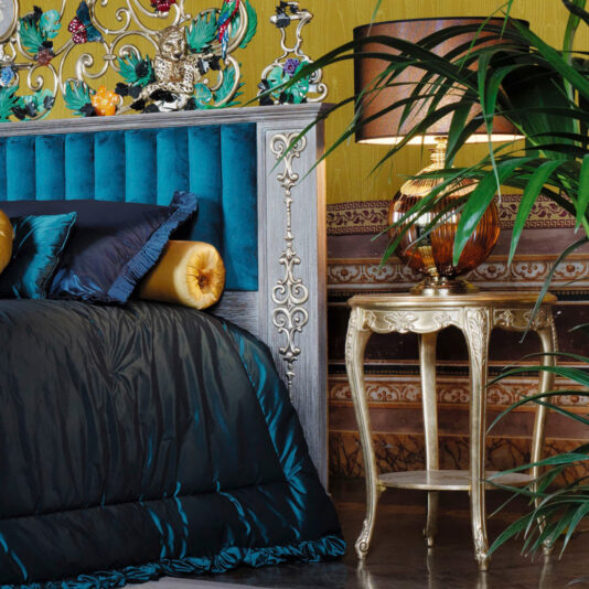 A luxurious bedroom with an ornate headboard featuring blue tufted upholstery and metallic accents. The bed is dressed in rich, dark bedding with golden pillows. Beside it sits a **Rococo Style Round Gold Bedside Table** holding a vintage lamp with a golden base and an amber shade. A large green plant adds a touch of nature to the scene.