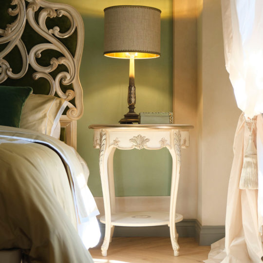 A bedroom featuring a vintage-style **Round Rococo Style Bedside Table** beside the bed. The table showcases ornate carvings and supports an elegant lamp with a beige shade. The bed boasts a green and white headboard adorned with creamy bedding, while a window draped with white curtains allows natural light to brighten the space.