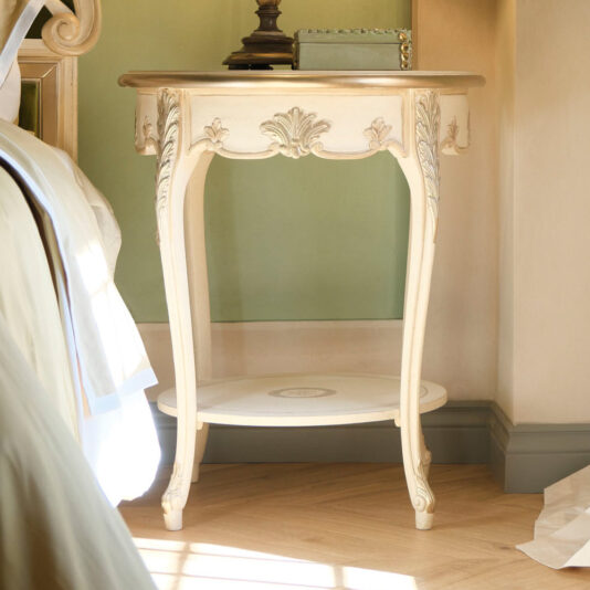 Round Rococo Style Bedside Table This image features the Round Rococo Style Bedside Table, showcasing its elegant, vintage-inspired design with a cream-colored finish. The bedside table boasts intricate carvings and curved legs, an ornate apron, and a small round shelf below. It is beautifully placed in a well-lit, cozy bedroom corner.