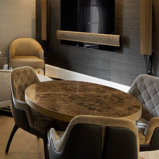 Small Modern Round Marble Dining Table A modern living room features a Small Modern Round Marble Dining Table surrounded by five cushioned chairs. A flat-screen TV is mounted on a dark accent wall with sconces on either side. An armchair and a sofa are arranged in the background over a sleek wooden floor, enhancing the contemporary vibe.