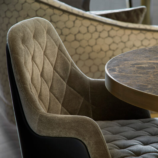 Small Modern Round Marble Dining Table A close-up of a modern, upholstered chair with a quilted pattern is positioned next to the Small Modern Round Marble Dining Table. Another chair featuring a geometric hexagon pattern can be seen in the background, creating a sophisticated interior setting.
