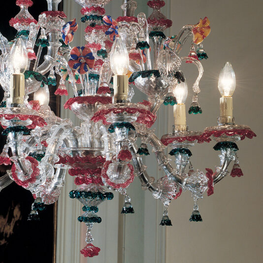 Statement Chandelier With Colourful Floral Detailing The Statement Chandelier With Colourful Floral Detailing boasts exquisite glasswork embellished with delicate leaf patterns in green, red, and clear. Several lit bulbs are interspersed among the vibrant glass decorations, highlighting the chandelier's intricate design and craftsmanship.