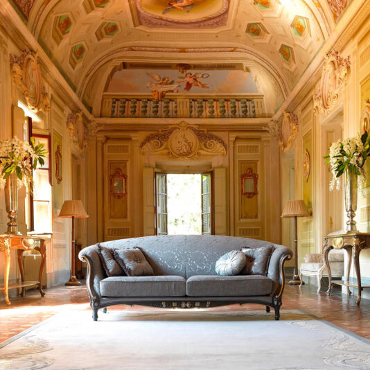 Classic Venetian Style Sofa An opulent, baroque-style room with ornate ceiling murals, tall windows, and decorative walls features a Classic Venetian Style Sofa in gray, which sits at the center on a light rug. The room is adorned with lavish furniture, floor lamps, and large floral arrangements on each side. Soft light filters in.