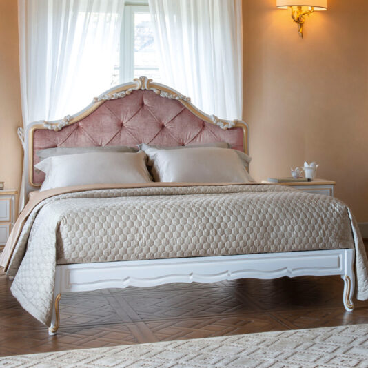 A luxurious bedroom features the Classic Button Upholstered Bed, a king-sized piece with a tufted pink velvet headboard and an elegant white frame. The bed is adorned with a beige quilted bedspread and matching pillows. Light floods the room through sheer white curtains, illuminating the warm decor.