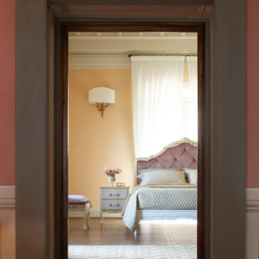 A cozy bedroom is seen through an open doorway. The room features a Classic Button Upholstered Bed, adorned with pillows and a quilted bedspread. A small bedside table with drawers holds a vase of flowers. Soft curtains hang over a tall window, letting in natural light.