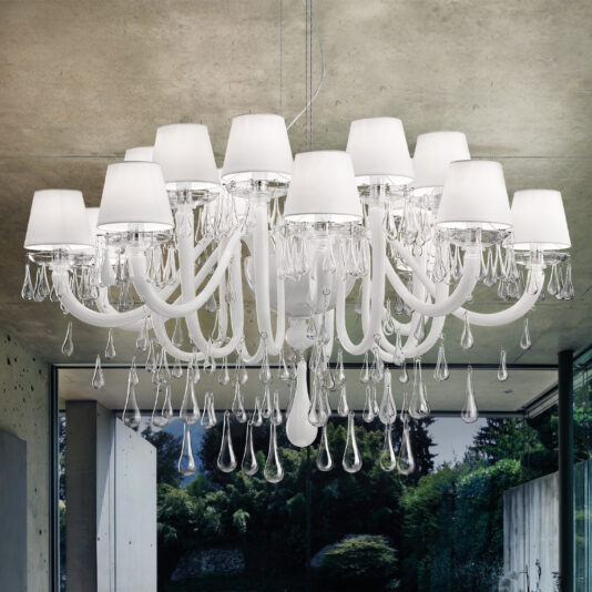 A Classic Glass Teardrop Accent Chandelier, featuring multiple white lampshades and exuding elegance, hangs prominently from the ceiling in a modern, minimalistic room. The backdrop consists of large windows that reveal a greenery-filled outdoor view.