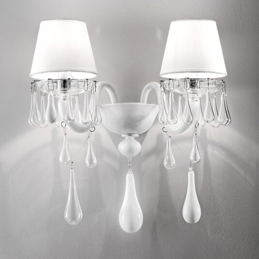 The Classic Glass Teardrop Accent Wall Light is a double wall sconce adorned with white lampshades and elegant, teardrop-shaped glass pendants hanging from each side. It boasts an ornate central fixture with a smooth glossy finish, mounted on a white wall, casting a warm and stylish glow.