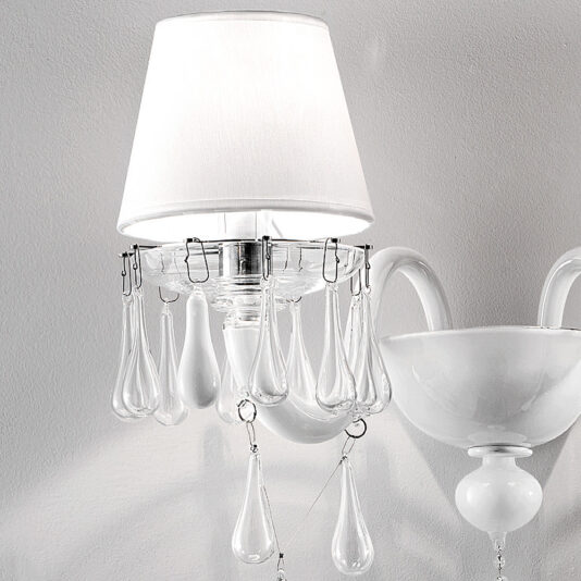 Classic Glass Teardrop Accent Wall Light A white wall-mounted lamp with a fabric shade and decorative hanging glass droplets stands beside the Classic Glass Teardrop Accent Wall Light, featuring a round base and curved arm. The lamp emits a soft light, creating subtle shadows as it reflects off the glass.