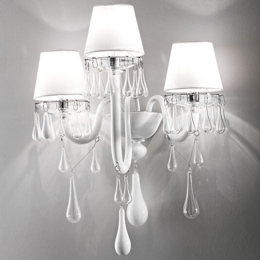 The Classic Glass Teardrop Accent Wall Light With Three Arms features three white lampshades, each adorned with clear teardrop-shaped crystal pendants. Mounted on a white base with curved arms, this elegant fixture adds a touch of sophistication to the decor against a light-colored wall.