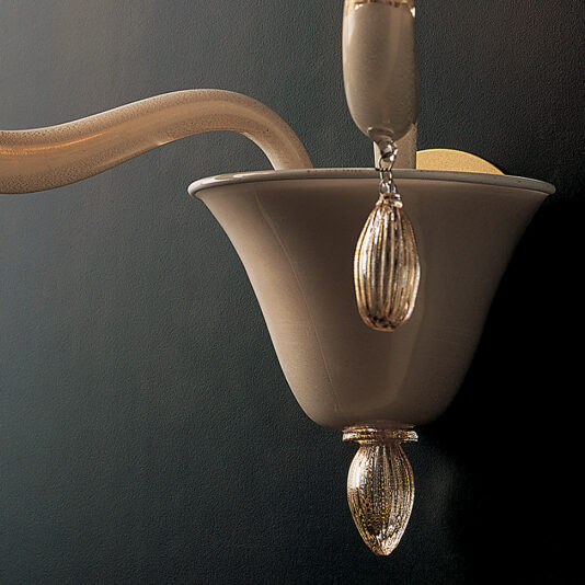 Classic Gold Glass Wall Light With Gold Frame Close-up of the Classic Gold Glass Wall Light With Gold Frame, showcasing its elegant and timeless design. The fixture features a flared, trumpet-shaped structure with a metallic finish, complemented by decorative glass elements that dangle from the base. Mounted on a dark textured wall, it casts subtle shadows.