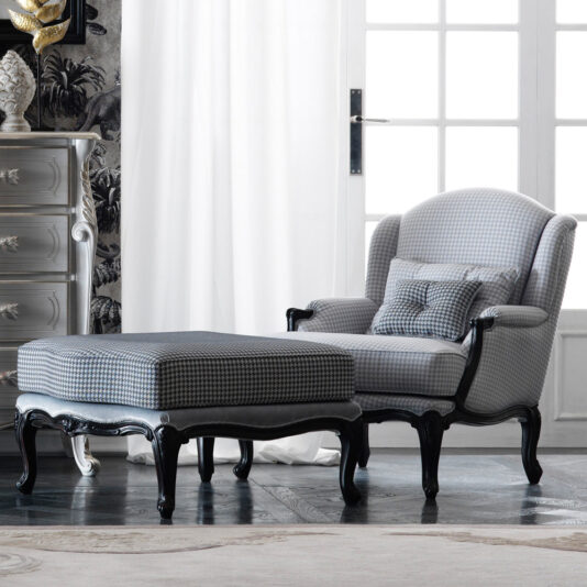 A sophisticated living room corner featuring the Classic Louis Style Armchair and Footstool, both upholstered in a houndstooth pattern. These pieces are supported by dark wooden legs adorned with intricate carvings, and are positioned near white curtains and a French door, with a chest of drawers to the side.