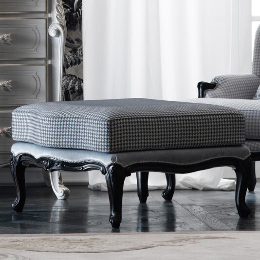 A square ottoman with a houndstooth patterned cushion and black wooden legs stands on dark hardwood flooring. Positioned in front of the Classic Louis Style Armchair And Footstool and beside a tall chest of drawers, the room is adorned with white curtains and a patterned rug, creating an elegant ambiance.
