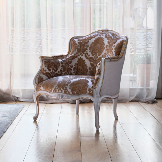 The Classic Louis Style Occasional Chair, an elegantly upholstered piece with a white wooden frame and floral-patterned fabric in warm shades of brown and gold, is positioned on a wooden floor near large sheer curtains that let soft, natural light fill the room—ideal as an occasional chair.