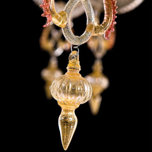 Classic Ornate Gold And Red Glass Chandelier A close-up of the Classic Ornate Gold And Red Glass Chandelier showcases its intricate ribbed detailing and pointed end, with multiple similar golden and red glass ornaments hanging from it. The background features a black backdrop with more of these beautiful ornaments visible in a blurred effect.