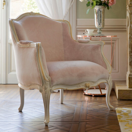 The Classic Style Louis Armchair and Footstool, adorned in a plush pastel pink fabric and set within an elegant white ornate wooden frame, rests on a patterned wooden floor. A side table behind it features a polished metal vase filled with flowers. Natural light flows through a window draped with white curtains in the background, enhancing the classic style ambiance.