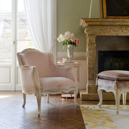 A luxurious room features the Classic Style Louis Armchair and Footstool upholstered in light pink velvet with ornate white frames. The furnishings are set on a patterned wooden floor near a grand stone fireplace, and a vase of pink and white roses sits on a side table beside the armchair.