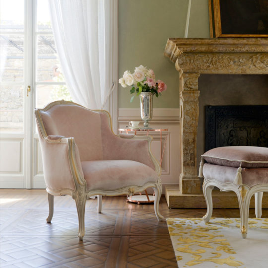 A luxurious room features the Classic Style Louis Armchair and Footstool upholstered in light pink velvet with ornate white frames. The furnishings are set on a patterned wooden floor near a grand stone fireplace, and a vase of pink and white roses sits on a side table beside the armchair.