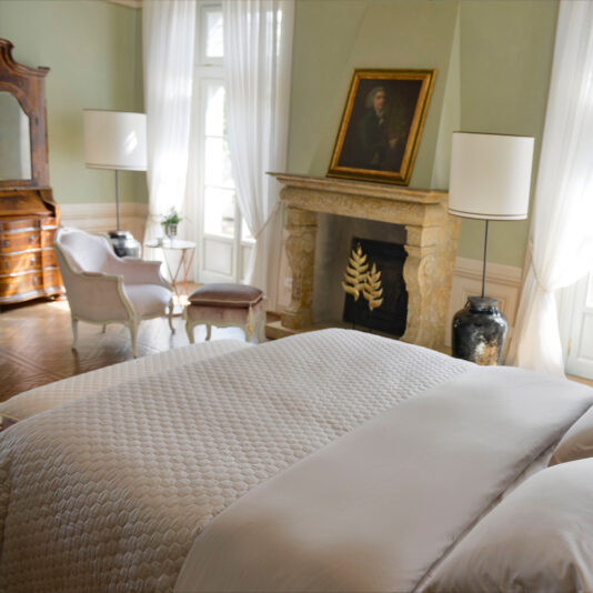 A serene bedroom with a cozy double bed adorned with a white quilted comforter, a fireplace with a framed portrait above it, and two Classic Style Louis Armchairs And Footstools beside a small table in front of a sunlit window. Elegant lamps and a wooden dresser add classic charm to the room.