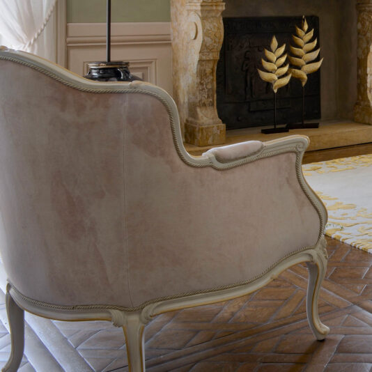 Beige Classic Style Louis Armchair And Footstool Featuring Carved Wooden Trim. In the background, a stone fireplace holds two gold leaf sculptures. Tuscan Rose Rugs’ Light-Patterned Carpet.