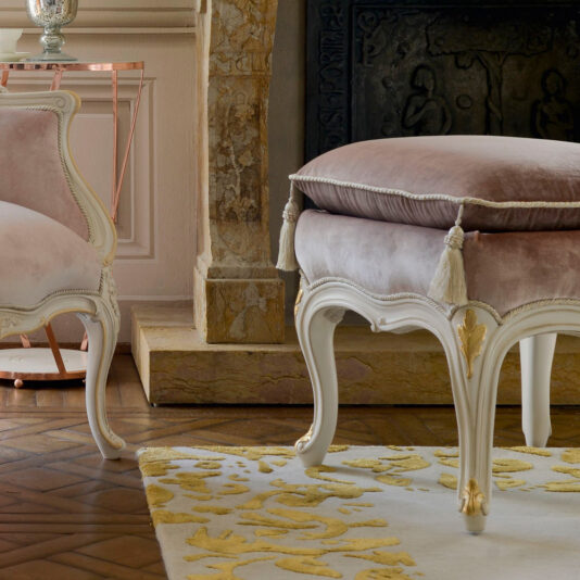 A close-up of an elegant interior featuring a plush cushioned stool with beige upholstery and gold accents, placed on a white and gold patterned rug. The Classic Style Louis Armchair And Footstool are visible in the background alongside a fireplace adorned with a decorative screen.