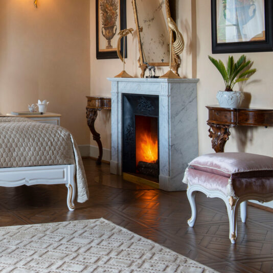 A cozy, elegant bedroom showcases a lit fireplace with a marble surround, a bed dressed in a beige quilted coverlet, and two wooden side tables flanking the white and gold carved bench. A Classic Style Louis armchair and its matching footstool rest beside the plush rug that partially covers the polished wooden floor. The walls are adorned with classic paintings, completing the sophisticated look.