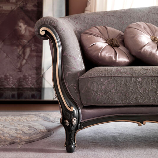 Classic Venetian Style Sofa The Classic Venetian Style Sofa boasts intricate, dark wooden armrests and floral-patterned upholstery, complemented by two round, tufted pillows. This elegant piece is perfectly showcased in a sophisticated setting featuring a textured carpet and an art piece visible in the background.