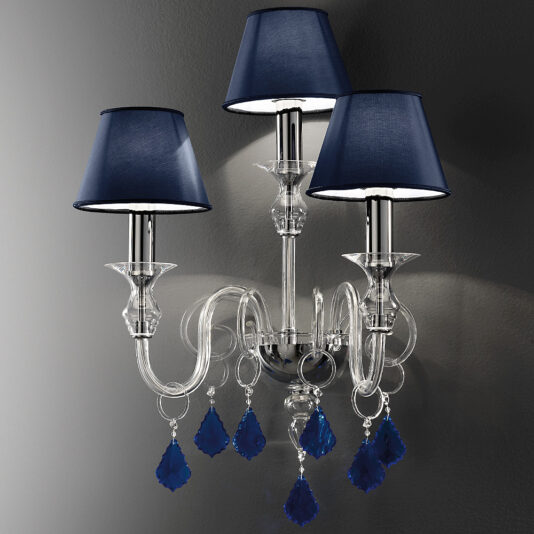 The Classic Wall Light With Blue Crystal Drops features a polished chrome finish and an ornate design. Each of its three arms holds a light adorned with navy blue lampshades, while elegant blue crystal drops hang gracefully from the arms. The sconce is tastefully mounted on a plain dark background, accentuating its sophisticated aesthetic.