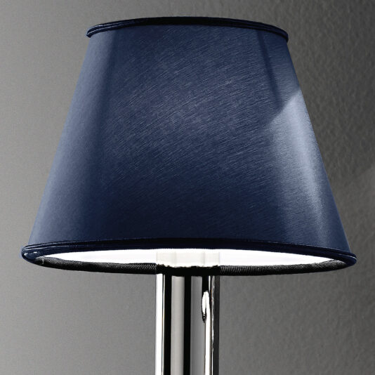 Classic Wall Light With Blue Crystal Drops A close-up of a table lamp featuring a sleek, shiny silver base paired with a dark blue, slightly tapered fabric lampshade. The background displays a simple, textured gray wall. The illuminated shade emits a gentle light that evokes the elegance of the Classic Wall Light With Blue Crystal Drops.