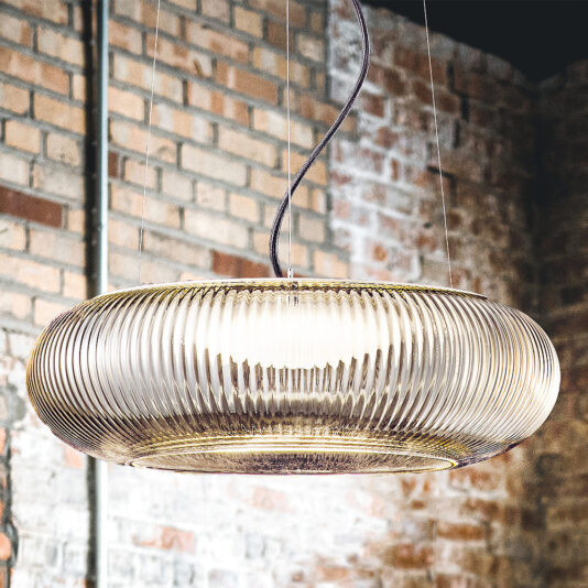 The Contemporary Ribbed Glass Pendant Light With Gold Frame hangs from the ceiling in a room with exposed brick walls. Its circular glass design features vertical ridges, offering a modern and elegant appearance. The pendant is suspended by a black cord, enhancing the industrial look of the space.