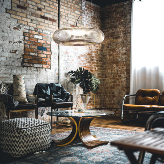 A stylish living room with exposed brick walls features a Contemporary Ribbed Glass Pendant Light With Gold Frame hanging from the ceiling. The space includes a glass coffee table with a unique wooden base, a patterned ottoman, a leather chair, and potted plants, all illuminated by natural light streaming through the window.