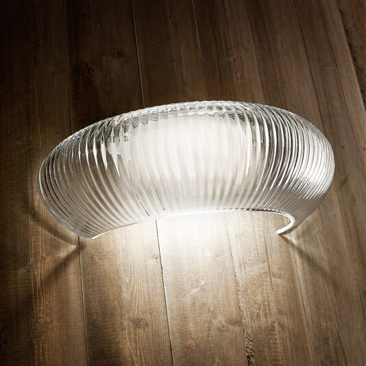 The Contemporary Ribbed Glass Wall Light, with its curved design, casts warm illumination onto a wooden wall, creating a soft glow that highlights the texture of the wood and the intricate details of the glass.