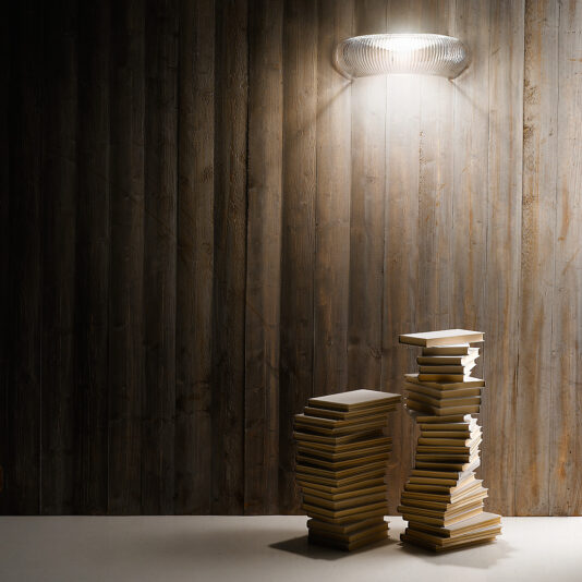 Contemporary Ribbed Glass Wall Light A modern, minimalist room features a textured wooden wall beautifully illuminated by the sleek Contemporary Ribbed Glass Wall Light. Two tall, irregularly stacked piles of books rest against the wall on a plain white floor, creating a serene and contemporary ambiance.