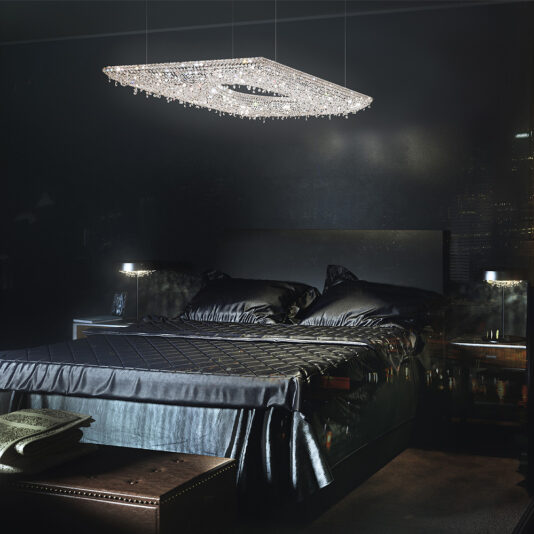 A luxurious bedroom featuring a dark, elegant design with a large, black bed covered in satin bedding. The Contemporary Suspended Crystal Pendant Chandelier hangs above the bed, casting a soft glow. Two small, modern wall-mounted lamps flank the headboard, and a trunk sits at the foot of the bed.