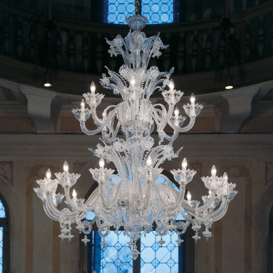 A Deluxe Floral Clear Glass Chandelier, adorned with intricate clear glass designs and multiple lit candles, hangs from the ceiling in a grand hall. The backdrop features arched windows with decorative panes, allowing natural light to complement the chandelier's glow.