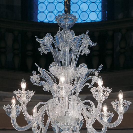 Deluxe Floral Clear Glass Chandelier The Deluxe Floral Clear Glass Chandelier is an intricate, multi-tiered masterpiece adorned with detailed floral and leaf designs. Each of its many lights is enclosed in delicate clear glass flowers, beautifully illuminating the ornate decor. The background highlights an architectural element with circular window designs.