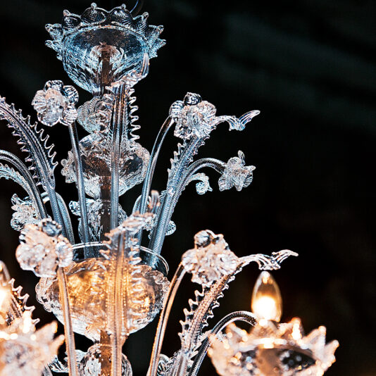 Deluxe Floral Clear Glass Chandelier A close-up of the Deluxe Floral Clear Glass Chandelier showcases its classic design with intricate glass details. The ornate fixture features delicate floral elements, clear glass flowers, and leaves, with a glowing bulb visible. The meticulous craftsmanship highlights fine detailing and elegance, reflecting light in various directions.