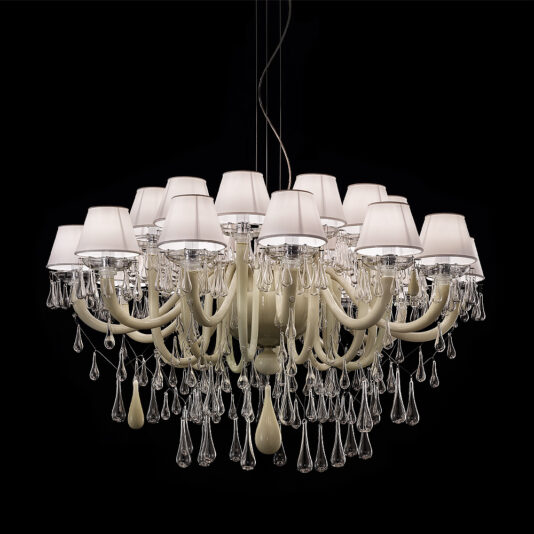 The Deluxe Glass Teardrop Accent Chandelier boasts multiple white lampshades perched atop elegant curving arms. Beneath the shades, glass teardrop accents dangle, creating a sparkling effect. The chandelier is set against a black background, highlighting its intricate design and opulence.