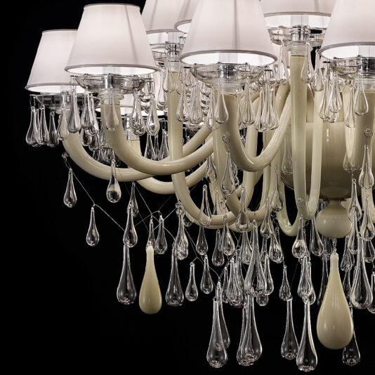 Deluxe Glass Teardrop Accent Chandelier The Deluxe Glass Teardrop Accent Chandelier is an elegant piece with ornate beige arms and white lampshades. It boasts numerous dangling glass teardrops, creating a luxurious and intricate design. The chandelier's exquisite details are further enhanced by the black background.