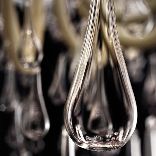 Deluxe Glass Teardrop Accent Chandelier Close-up of a Deluxe Glass Teardrop Accent Chandelier, showcasing an intricate play of reflections and refractions. The background is blurred, revealing other similar glass pieces and a softly focused light source. The overall tone exudes elegance and sophistication.