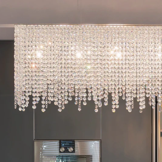 Deluxe Modern Narrow Crystal Chandelier The Deluxe Modern Narrow Crystal Chandelier emits a sparkling glow with cascading crystal tiers, creating an elegant and luxurious atmosphere against a backdrop of gray cabinetry and a cooking range.