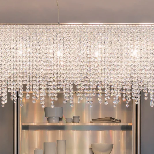 Deluxe Modern Narrow Crystal Chandelier The Deluxe Modern Narrow Crystal Chandelier elegantly illuminates the kitchen with its cascading crystal strands. Below, sleek open shelving highlights white dishes, cups, and a bowl.