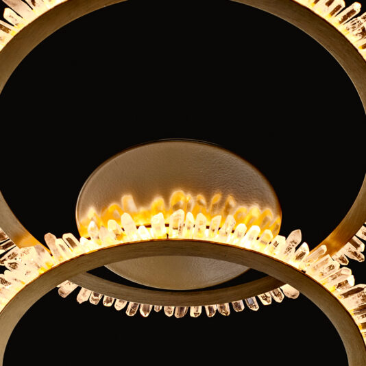 Double Ring Quartz Crystal Gold Ceiling Light A close-up view of the elegant Double Ring Quartz Crystal Gold Ceiling Light features concentric circular metal rings adorned with quartz crystal accents. This gold ceiling light emits a warm, glowing radiance against a dark background, highlighting the intricate details of its design.