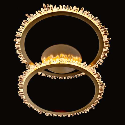The Double Ring Quartz Crystal Gold Ceiling Light features a contemporary chandelier design with two circular, gold rings embellished with quartz crystal accents. Its double-ring configuration emits a warm, ambient light against a black background, while the crystals catch and reflect the light to produce a sparkling effect for an elegant ceiling lighting solution.