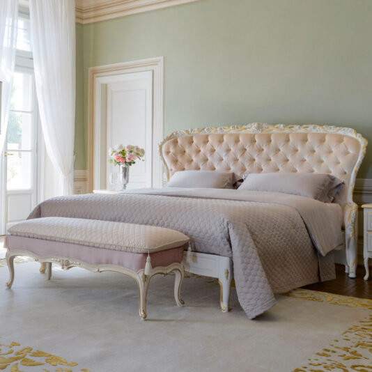 Luxurious bedroom featuring the Elegant Button Upholstered Winged Bed adorned with gray bedding and complemented by a matching bench at the foot. The room boasts tall white doors, green walls, ornate molding, and a vase of flowers on a side table. Natural light streams through sheer curtains.